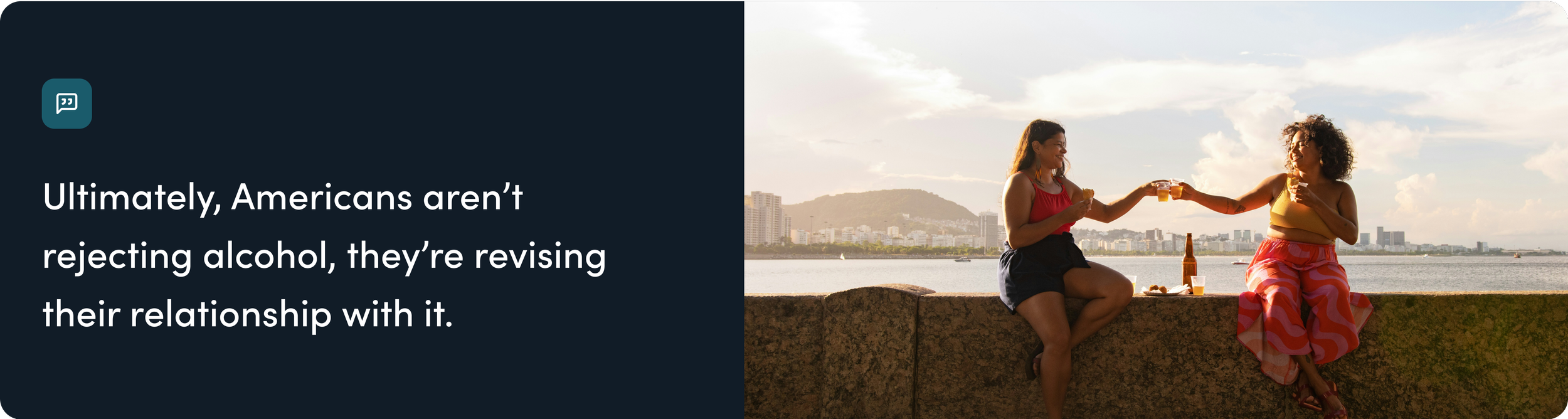 Two women sitting on a stone wall, clinking glasses of beer, with a city skyline and river in the background during sunset.